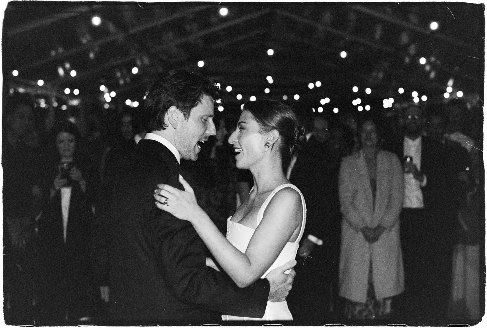 A first dance under festoon lights with guests around, a black and white film frame