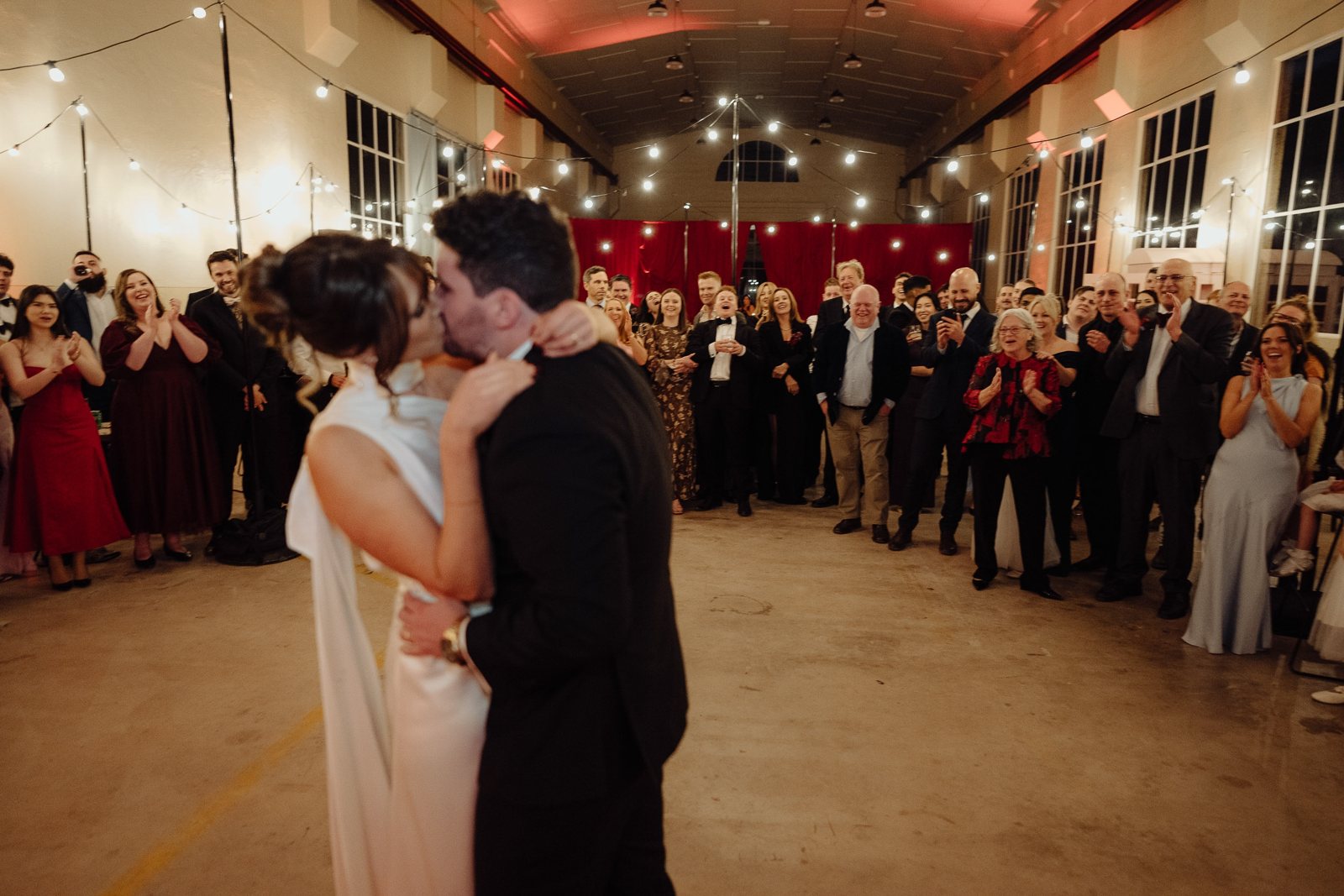 A first dance in a hall lit with string lights, guests gathered around the couple