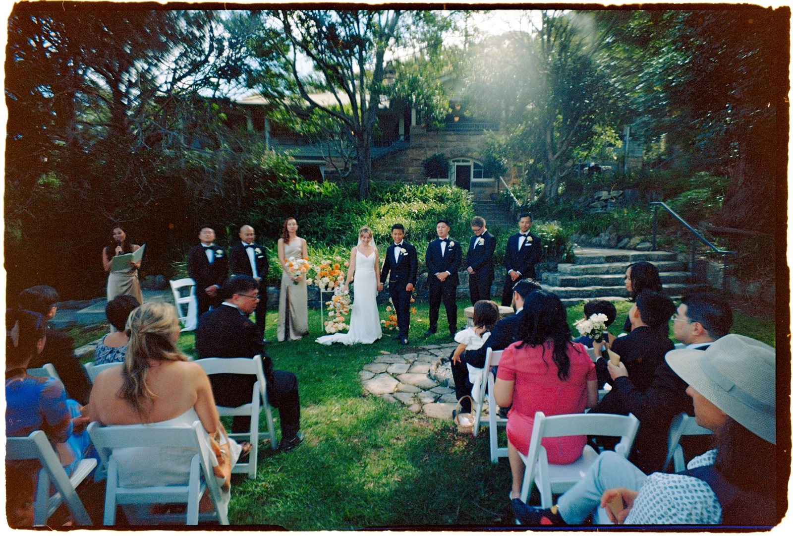 A garden ceremony under tall trees, guests seated in the shade, a colour film frame