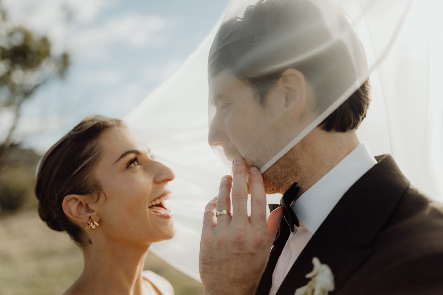 A bride laughing under a veil held over her and her groom in warm evening light
